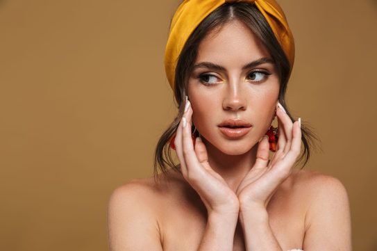 Close up beauty portrait of an attractive young topless woman wearing headband standing isolated over brown background, posing, touching her face, looking away